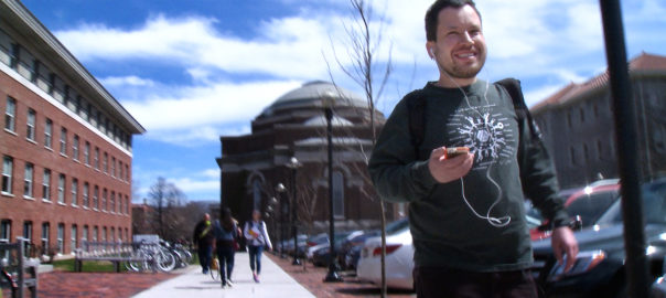 A man stands on a sidewalk, smiling with headphones in.