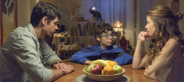 A young boy in a blue cape sits at the head of a table. A man and a woman sit on either side of the table looking at him.