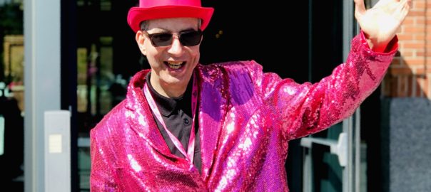 A man in a shiny pink hat and suit jacket stands outside the Prospect theater with his arms in the air