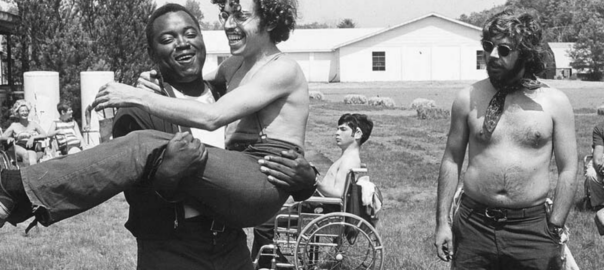 A man carries another man in his arms while a third man stands to the right of them and looks at them. A person in a wheelchair is in the background.