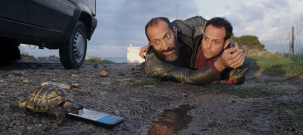 Two men lay on the ground wrestling while a turtle moves closer to a cellphone on the ground
