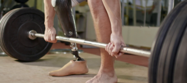 Hands grasp a barbell on the group. One of the weightlifter's legs is a prosthetic.