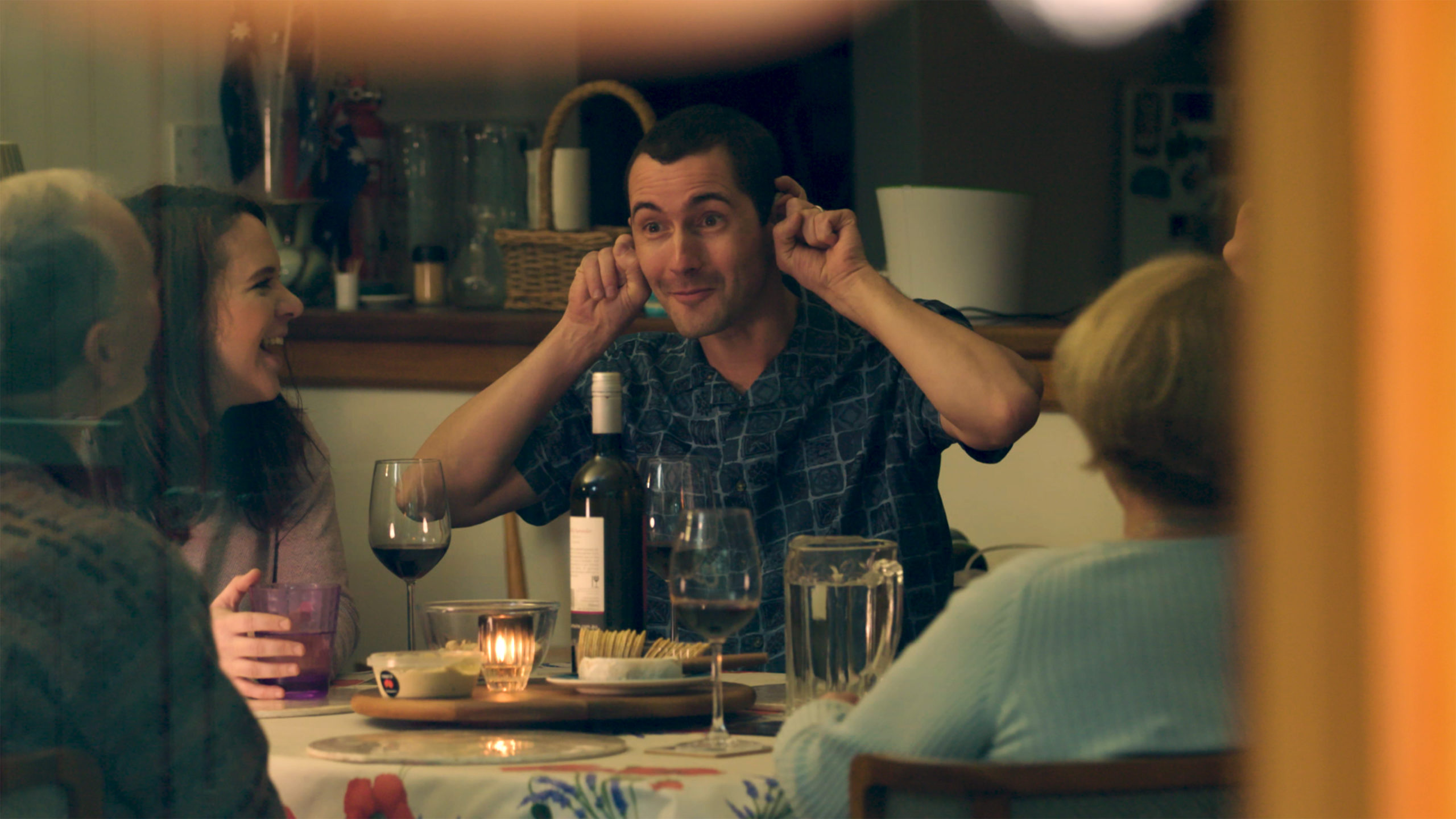 A man pulls on his ears comically at a dinner table surrounded by several other people