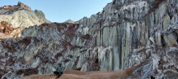 A man sits on a pile of red earth with a large rock formation behind him