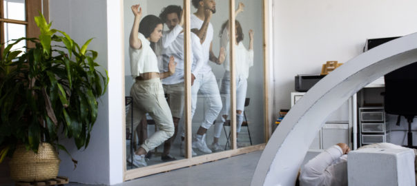 A group of dancers dressed in white lean against a wood-framed glass wall in an industrial space