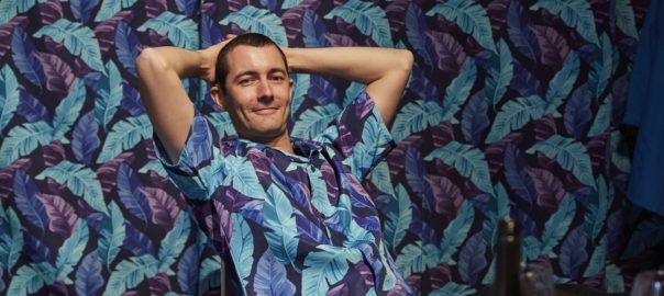 A man in a blue and purple print shirt leans back in a chair with his hands laced behind his head. His shirt matches the wallpaper behind him exactly.