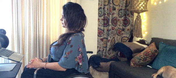 A woman in a wheelchair looks at a laptop on a desk. There is a grey couch behind her.