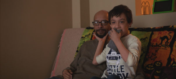 A father and son sit on a couch together. The boy holds his father chin in his hand.