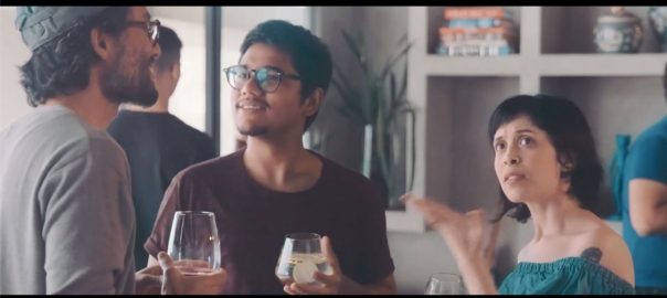 Two young men and a woman stand together talking and signing. The two men both have wine glasses in hand.