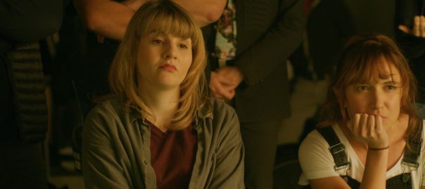 A woman in a wheelchair, wearing a green sweater, sits next to a woman in overalls on a film set