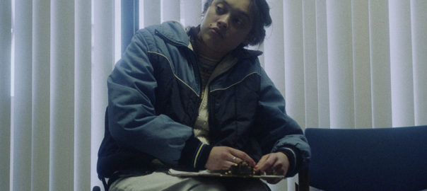 A teenage girl, who is a wheelchair user, in a puffy coat holds a clipboard in a waiting room.