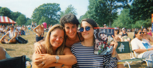 Three women sit together on a grassy field with other groups of people around them. One of the women holds up an issue of Curve magazine