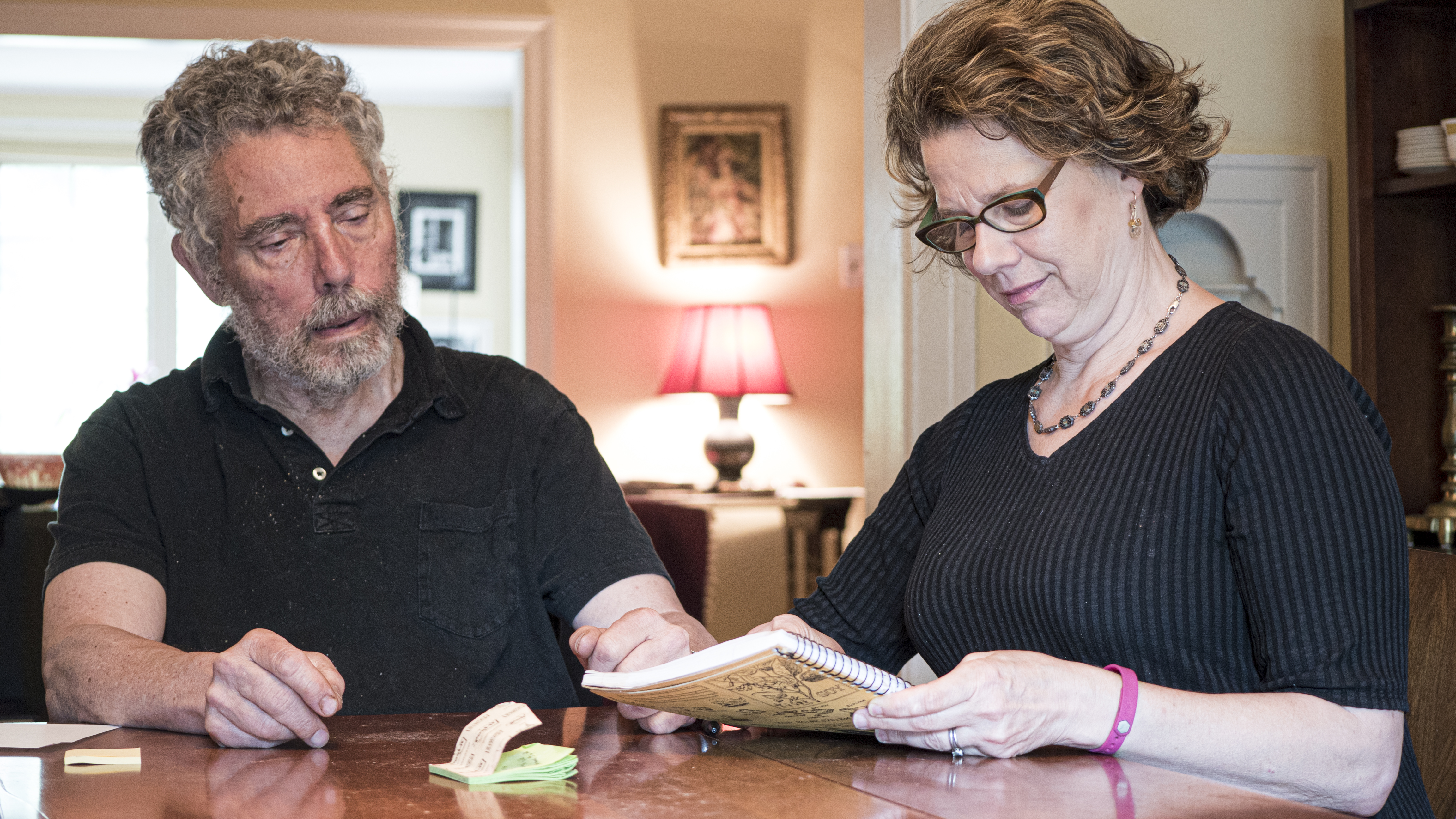 A man and a woman sit at a table together. They are looking over a notebook the woman holds in her hands.