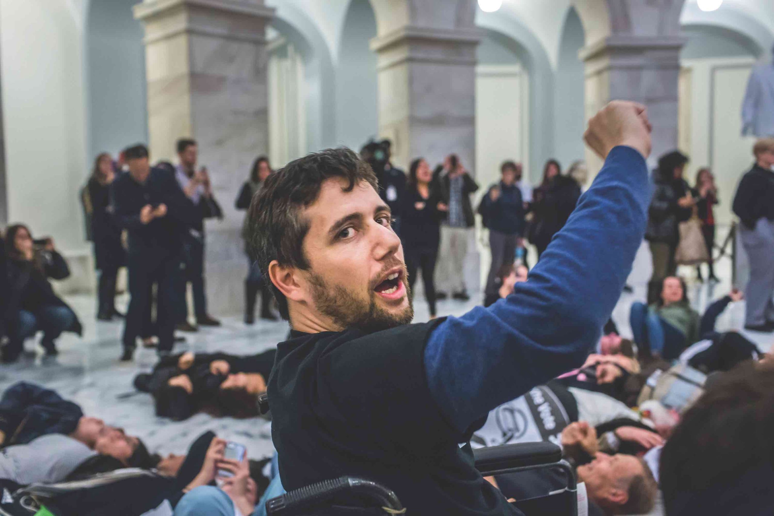 A man in a wheelchair raises one fist in the air. Behind him, several protestors lay on the floor while others look on around them.