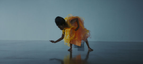 A woman in a scenery of blue bows down into a dance posture.