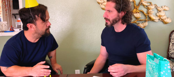 Two brothers, one a little person, sit at a table facing each other with yellow birthday cones on their heads.