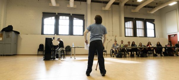 A person with a walking stick faces a group in a rehearsal room, one person at the piano and actors with various disabilities seated at a table.