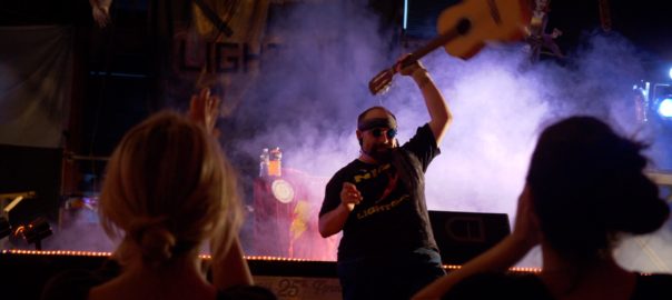 Standing in front of a stage illuminated by blue fog, a man proudly holds up a guitar.