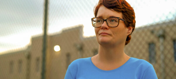 a short-cut redhead woman wearing glasses and a light blue t-shirt standing in front of a fenced structure.