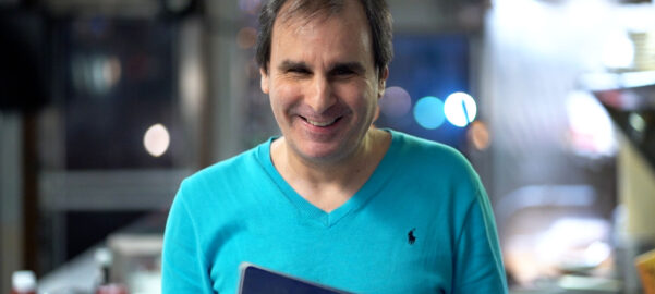 a smiling man wearing a blue v-neck shirt, holding a menu