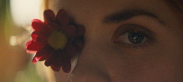 the upper part of a young woman's face, staring at the viewer, with a flower covering one of her eyes