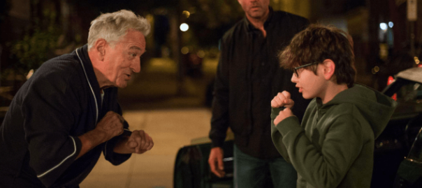 A man stands by and watches as an older smiling man and a little boy clench their fists in fighting position.