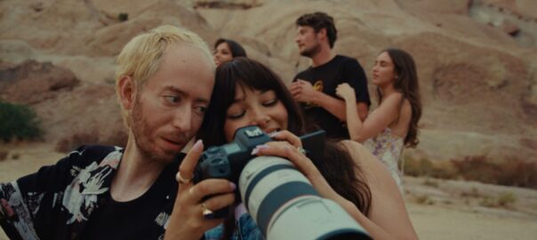 A young man and woman smile and look through a camera, with young adults in the background.