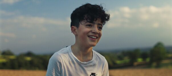 A young boy stands in a field, smiling at the horizon off camera.