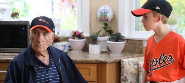 A boy in an Orioles baseball cap and team uniform stands next to an old man sitting at a kitchen table wearing the same team cap.