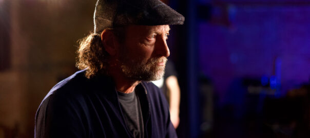 A man with trimmed facial hear and shoulder-length hair with a distinct cap looks into camera lights on a stage with a deep blue offstage backdrop.