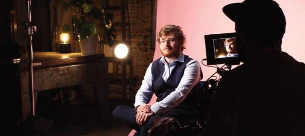 A man sits on a set with a pink backdrop as a videographer in the foreground stands with his back to us, filming the subject.