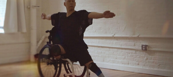 An Asian man in a baggy black outfit with a bald head strikes a pose in a dance studio with ivory walls. His eyes are closed and arms are outstretched.