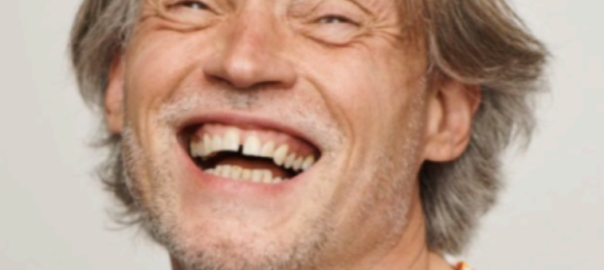 A headshot of Lawrence Carter-Long, a man with a full, cheerful smile, tilting his head a bit backwards, his brownish-grey hair covers his ears. He is wearing a light colored shirt with a pattern of orange flowers.