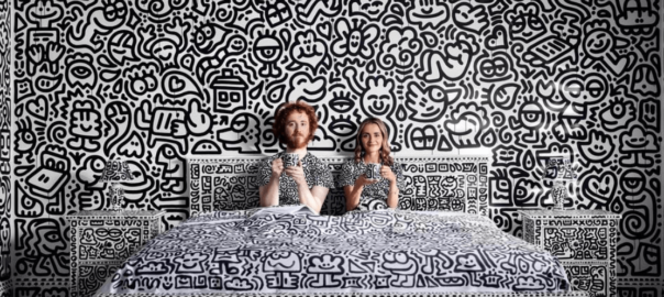 a bedroom entirely covered in black-and-white doodles, including the walls, furniture, bedding, and even the lamps. Two people sit on the bed, holding mugs and wearing clothing that blends into the doodle-covered background.